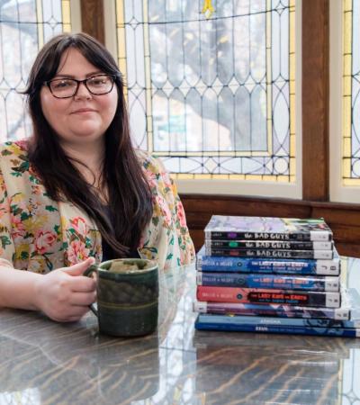 Photograph of Kelly Povero sitting at a table inside the library with a stack of books.