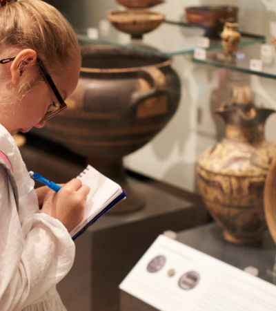 Photo shows a young student taking notes in front of artifacts in a museum case