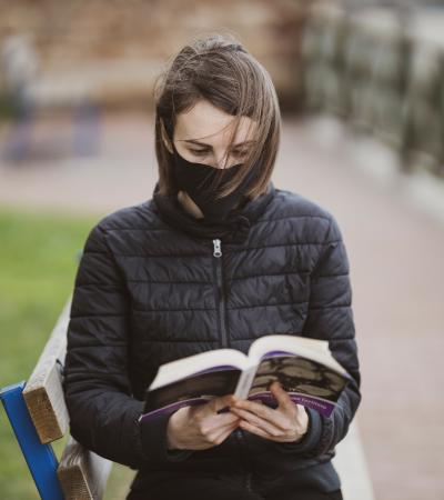 A woman wearing a mask reading on a park bench