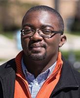 man with glasses looking at camera wearing a blue striped collared shirt, orange sweater, and black jacket