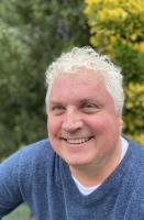  Headshot of man with curly white hair smiling at camera and wearing a blue sweater with a white shirt underneath.