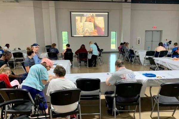 Photograph of a large room with people sitting at tables facing a projector screen. Jolene Poore is on the large screen reading from a book.