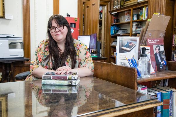 Photograph of Kelly Povero sitting at the circulation desk with a stack of books.