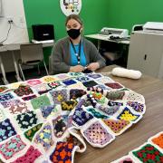 Photograph  of a masked person sitting at a table crocheting the blanket