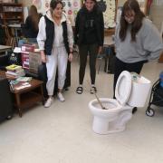 Photograph of three students in a classroom looking at a toilet with a plunger