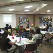 Photograph of room full of participants sitting at tables. They are looking at a slideshow.