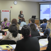 Photograph of room full of participants sitting at tables. They are looking at a slideshow with a presenter. 