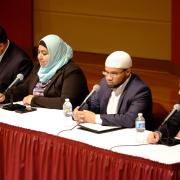 Four panelists sit side by side across a long table with a red tablecloth.