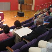 Attendees are seated in an auditorium with a seated woman addressing the crowd.