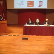 A woman standing in front of a podium addresses the four panelists seated at a long table with a red tablecloth.