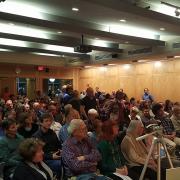 A large crowd of people are seated while a line forms with attendees waiting to ask questions.