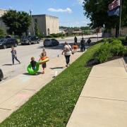 Photograph shows children outside playing a sidewalk chalk game with pool floaties on.