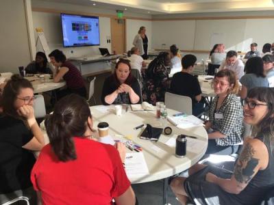 People sit in a group around a table with notebooks and pens in front of them in a large conference room.