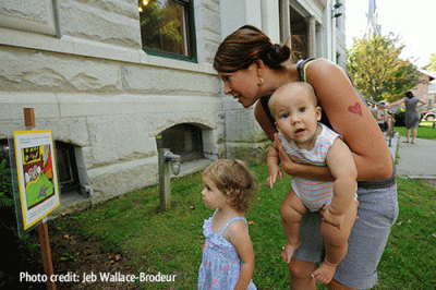 A woman holding a baby and reading a StoryWalk post with a toddler